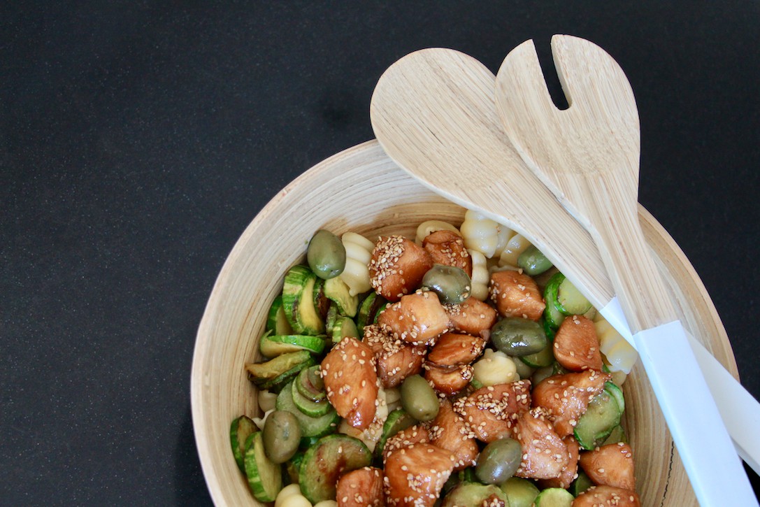 Wooden bowl filled with pasta salad featuring chicken, zucchini, and olives, in a simple décor