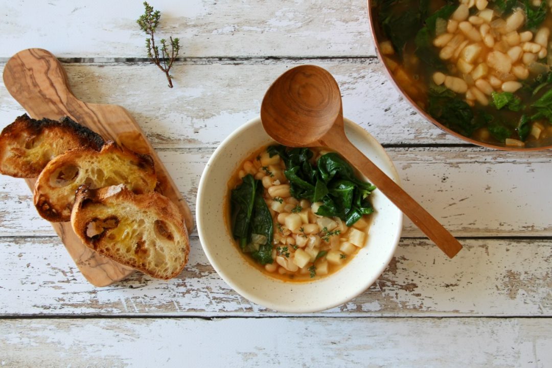 Rustic Tuscan white bean soup with wilted spinach, served in a speckled ceramic bowl with toasted bread on a wooden board