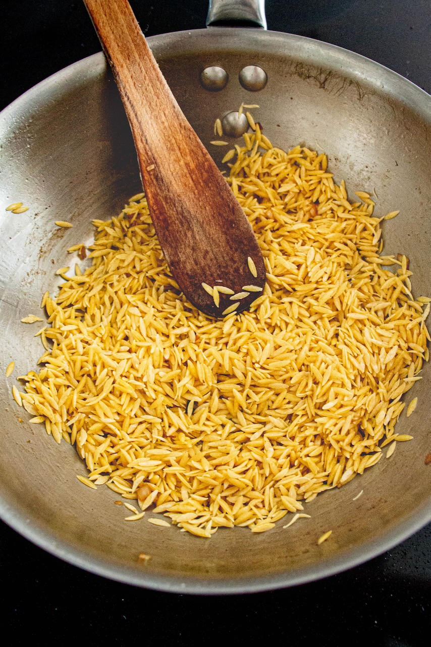 Golden orzo in a skillet with a wooden spoon, preparing the orzo recipe