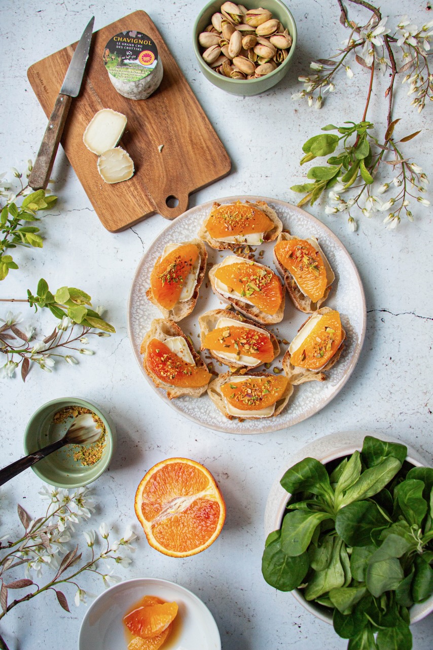 Plate of bruschettas with Crottin de Chavignol, orange segments, and pistachios, viewed from above