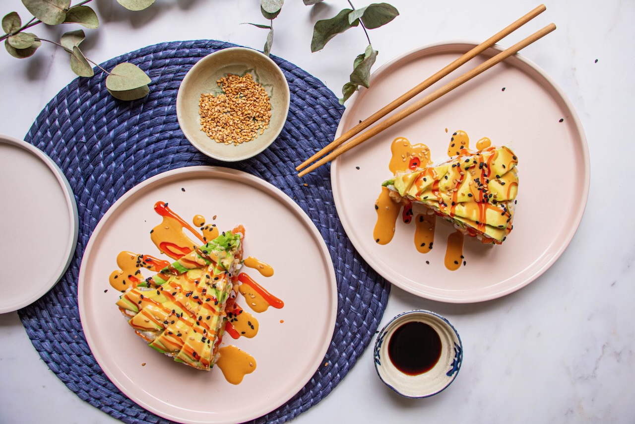 Two slices of Avocado Salmon Sushi Cake on pink plates, with sauce and sesame seeds