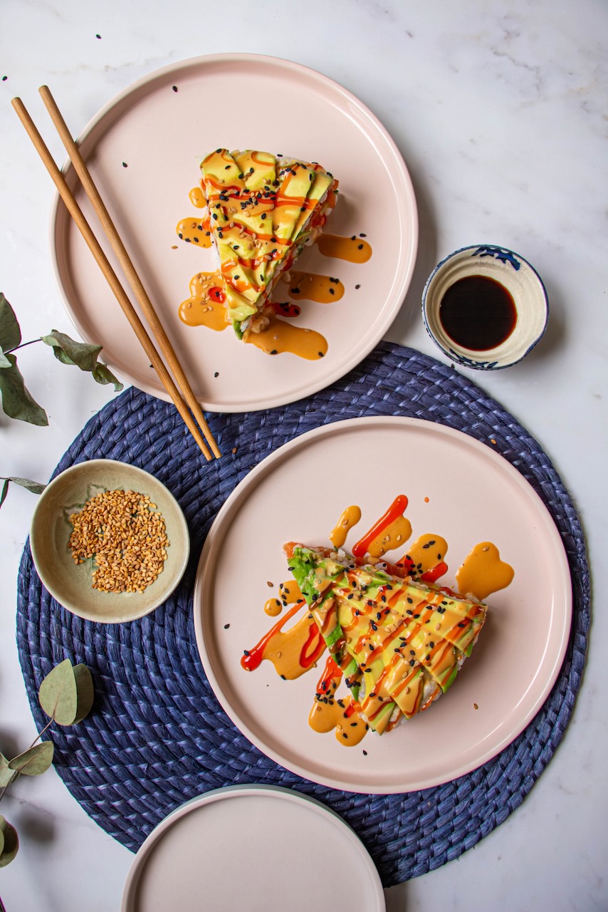 Two servings of Avocado Salmon Sushi Cake on pink plates, drizzled with colorful sauces