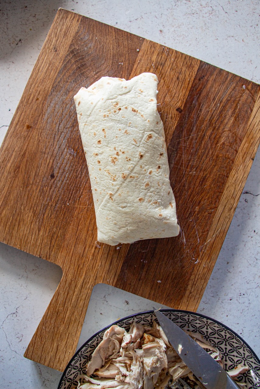 Rolled wrap on a wooden board, with pieces of chicken on a plate alongside